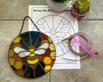 May include: A stained glass honey bee with a honeycomb pattern, a glass cutter, and a roll of tape on a white paper pattern with numbers and the text "Honey Bee Piece Numbered".