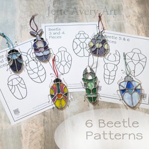 Può includere: Sono esposti sei ornamenti a forma di scarabeo in vetro colorato di vari colori, tra cui blu, verde, viola e giallo. Ogni scarabeo ha un design diverso ed è sospeso da un filo. L'immagine mostra anche modelli con il testo "Beetle Patterns."