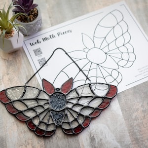 May include: A stained glass moth suncatcher with red, white, and black glass pieces. The moth is hanging from a black chain and is on a wooden surface. The image also includes a printed sheet of paper with the words "Web Moth Pieces" and a black and white outline of a moth.