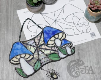 Op de afbeelding: Gebrandschilderd glaskunstwerk met blauwe paddenstoelkoppen, witte stelen en groene bladeren. Een spinnenbedel hangt eronder. Geplaatst op een grijze houten ondergrond met een paddenstoelpatroon en kleine potplanten.
