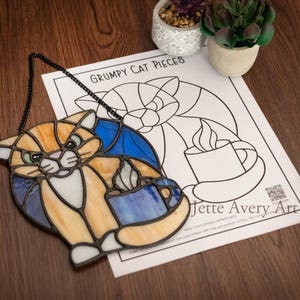 Peut inclure: Un chat en verre coloré tenant une tasse de café. Le chat est orange, bleu et blanc. L'image comprend le texte "Grumpy Cat Pieces" et "Jette Avery Art".