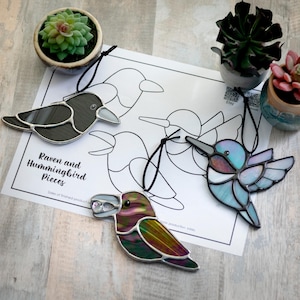 May include: Three stained glass bird ornaments, a black raven, a blue hummingbird, and a purple and green hummingbird, hanging from black cords. The ornaments are displayed on a white sheet of paper with a black and white outline of the birds. The paper has the text "Raven and Hummingbird Pieces" printed on it.