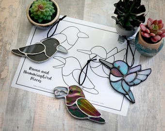 May include: Three stained glass bird ornaments, a black raven, a blue hummingbird, and a purple and green hummingbird, hanging from black cords. The ornaments are displayed on a white sheet of paper with a black and white outline of the birds. The paper has the text "Raven and Hummingbird Pieces" printed on it.