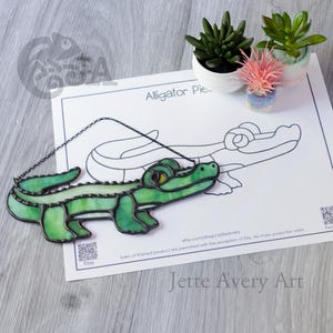 Może przedstawiać: Zielony witrażowy aligator z żółtymi oczami wisi na łańcuchu. Aligator jest umieszczony na rysunku aligatora z tekstem "Alligator Pie". Małe rośliny doniczkowe znajdują się w tle.