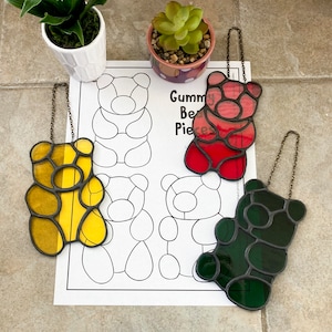 May include: A set of four stained glass gummy bear pieces. The bears are yellow, red, green, and black. The bears are hanging from a string and are on a white background with a colouring page of gummy bears. The colouring page has the text "Gummy Bear Pieces".