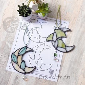 May include: Two crescent moon suncatchers made of stained glass with dark metal soldering. One features pale blue and cream glass with a clear crystal bead, the other has green and white glass with a green and clear crystal bead, both hanging by cords.