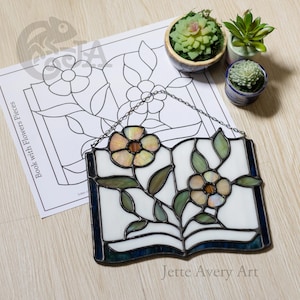 Puede incluir: Un atrapasoles de vidrieras con forma de libro abierto con dos flores, que presenta piezas de vidrio blanco, verde e iridiscente unidas por soldadura de metal oscuro. Se incluye un patrón de papel con la etiqueta Book with Flowers Pieces.