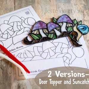 Op de afbeelding: Een gebrandschilderd glazen paddenstoelzonenvanger met paarse en witte paddenstoelen, groene bladeren en een blauwe swirl. De zonenvanger ligt op een wit vel papier met een zwart-witte omtrek van de zonenvanger.