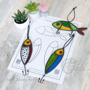 Pode incluir: Iscas de pesca em vitral colorido, vermelho, amarelo, verde e azul, com contornos e anzóis pretos. As iscas são exibidas em uma superfície branca com um desenho de peixe e o texto "Fishing Lures".