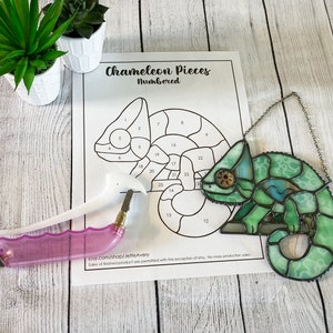 May include: A stained glass chameleon with a green, blue and white colour scheme. The chameleon is hanging from a silver chain. The image also shows a glass cutter and a numbered pattern for the stained glass project.