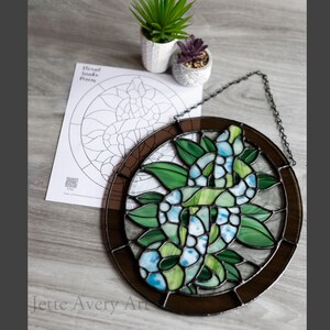 Pode incluir: Uma janela de vitral com uma cobra verde com escamas azuis e verdes, cercada por folhas verdes. A cobra está enrolada em um círculo e a janela é emoldurada em marrom.