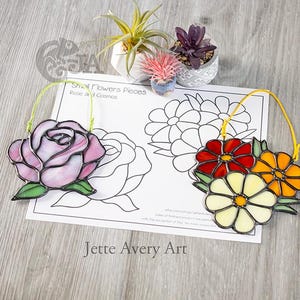 May include: Two stained glass flower ornaments: a pink rose and a cluster of red, yellow, and orange flowers. Each has a wire loop for hanging. The ornaments are on a paper with flower outlines and the text "Small Flowers Pieces Rose and Cosmos".