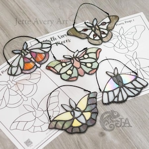 May include: A collection of stained glass moth ornaments in various colors, including orange, pink, green, and yellow. Each moth has a black wire loop for hanging. The image also shows a printed page with moth designs and the text "Jette Avery Art".
