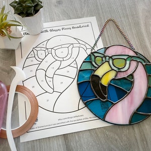 Puede incluir: Un flamenco rosa con gafas de sol de vidrio coloreado azul y verde. El flamenco es parte de un proyecto de arte de vitrales con piezas numeradas. La imagen también muestra herramientas y suministros para el arte del vitral, incluyendo cinta de cobre, un cortador de vidrio y una plantilla.