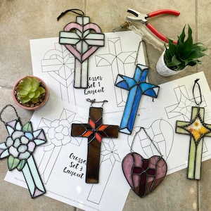 May include: A collection of stained glass crosses in various designs and colors, including pink, blue, brown, and green. Some crosses have floral or heart-shaped elements. The image also shows cross layout designs and small potted plants.