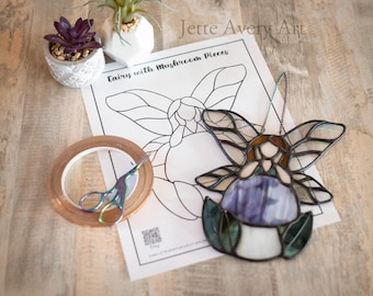 May include: A stained glass fairy with mushroom pieces, featuring a purple and white mushroom body, brown hair, and iridescent wings. The image includes a roll of copper tape, scissors, and a succulent plant.