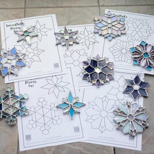 May include: A set of eight stained glass snowflake ornaments with a variety of colors and designs. The ornaments are displayed on top of printed sheets of paper with the title "Snowflake Pieces" and "Pieces Pg2".