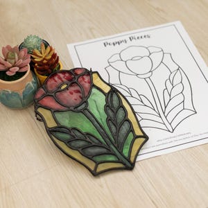 Peut inclure: Un vitrail repr&eacute;sentant un coquelicot rouge, vert et jaune, avec un contour noir. &Agrave; c&ocirc;t&eacute;, une page &agrave; colorier intitul&eacute;e "Poppy Pieces" et de petites plantes succulentes en pot.