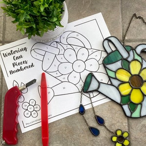 Puede incluir: Un atrapasoles de vidriera con forma de regadera con un diseño de flor amarilla y marrón, colgado de una cadena. Un patrón numerado para un proyecto de vidriera, un cortador de vidrio rojo y un rotulador rojo también son visibles.