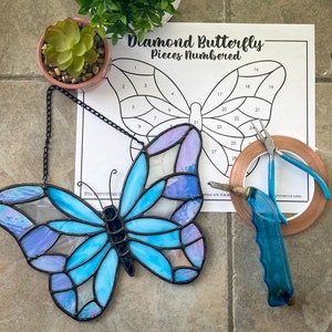 May include: A stained glass butterfly with blue and purple glass pieces. The butterfly is hanging from a black chain. The image also shows a printed sheet with the text "Diamond Butterfly - Pieces Numbered" and numbers 1 through 28. There are also tools for working with stained glass, including copper foil tape, a soldering iron, and a pair of pliers.