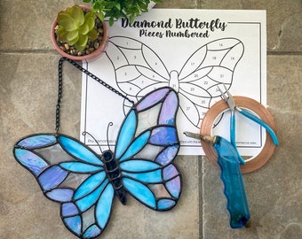 May include: A stained glass butterfly with blue and purple glass pieces. The butterfly is hanging from a black chain. The image also shows a printed sheet with the text "Diamond Butterfly - Pieces Numbered" and numbers 1 through 28. There are also tools for working with stained glass, including copper foil tape, a soldering iron, and a pair of pliers.