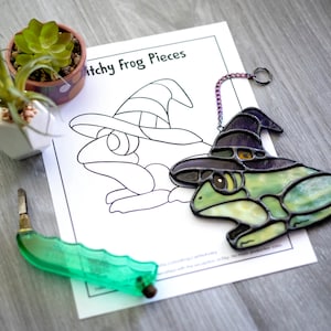 Könnte beinhalten: Eine Glasmalerei-Frosch mit einem Hexenhut, in grün, gelb und schwarz. Der Frosch sitzt auf einem weißen Blatt Papier mit dem Aufdruck "Itchy Frog Pieces".