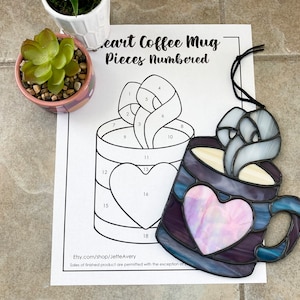 Puede incluir: Una taza de café de vidriera con un diseño de corazón, en tonos morados, azules y blancos. La taza tiene un centro en forma de corazón y un diseño similar a vapor en la parte superior. Un patrón numerado para la taza está al lado.