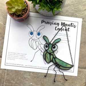 May include: A stained glass praying mantis pendant with a green and blue color scheme. The pendant is hanging from a black chain. The image includes a layout diagram for the pendant, which includes instructions for making the pendant.