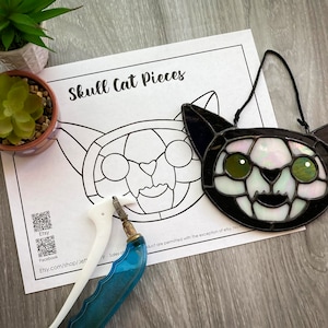 May include: A black and white stained glass skull cat with green eyes. The cat is hanging from a string and has a printed pattern on the glass. The cat is on a white sheet of paper with the words "Skull Cat Pieces" printed on it. There are also two QR codes on the paper.