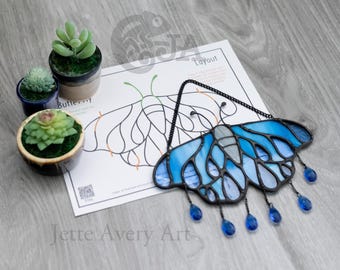 Op de afbeelding: Een blauw en grijs glas-in-lood vlinderornament, opgehangen aan een zwarte ketting. De vlinder heeft hangende blauwe glazen druppels. Een lay-out vel en kleine vetplanten in potten zijn ook zichtbaar.