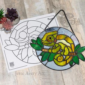 May include: Stained glass chameleon artwork in yellow, green, and brown, suspended by a black chain. The piece is set against a light blue backdrop and features green leaves. A black and white pattern is visible in the background.