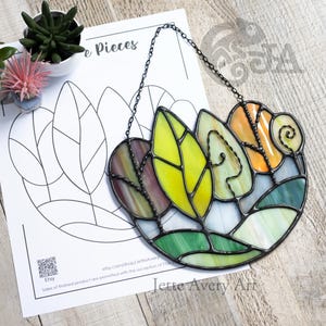 May include: A stained glass suncatcher featuring a collection of leaves and a spiral tendril in shades of yellow, orange, purple, and green, held together by dark metal solder lines and suspended by a black metal chain against a white paper pattern.