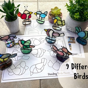 Peut inclure: Neuf figurines d'oiseaux en vitrail colorés avec des pattes et des antennes en fil de fer sont exposées. Les oiseaux sont de couleurs variées, dont le rouge, le bleu, le vert et le violet. Une feuille de papier avec des contours d'oiseaux et le texte "Standing Birds Pieces" est visible.