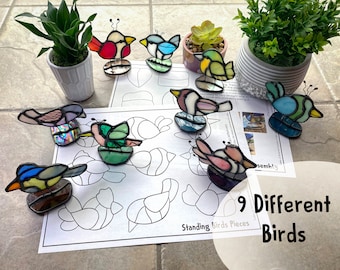 May include: Nine colorful stained glass bird figurines with wire legs and antennae are displayed on a white surface. The birds are various colors, including red, blue, green, and purple. A sheet of paper with bird outlines and the text "Standing Birds Pieces" is visible.