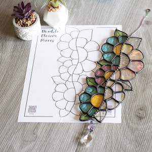 Könnte beinhalten: Ein Buntglas-Sonnenfänger mit einem Doppelblumenmuster in Blautönen, Rosa und Gelb. Der Sonnenfänger hängt an einer silbernen Kette und hat ein Kristallprisma, das an der Unterseite hängt. Der Sonnenfänger liegt auf einem bedruckten Blatt Papier mit den Worten "Double Flower Pieces" und einem QR-Code.