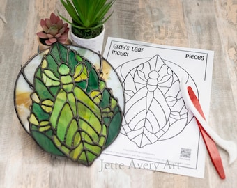 Op de afbeelding: Een kunstwerk van glas-in-lood dat een groene bidsprinkhaan op een blad voorstelt. Het stuk is omgeven door een zwarte lijst en wordt vergezeld door een zwart-witte omtrek van hetzelfde ontwerp, met de tekst "Gray's Leaf Insect" en "Pieces".