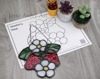 May include: A stained glass strawberry and flower suncatcher with red, white, and green colors. The suncatcher is hanging in front of a coloring page that says "Strawberry Pieces". Small potted succulents are in the background.