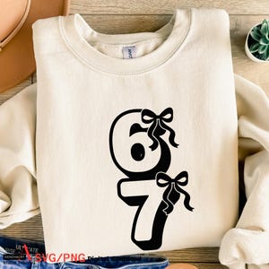 May include: Cream-colored sweatshirt with a black graphic of the numbers 6 and 7, each adorned with a bow. The sweatshirt is made of a soft material. The text "SVG/PNG Digital download" is visible at the bottom.