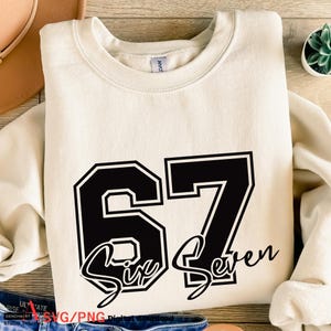 May include: Cream-colored sweatshirt with the number "67" in black, outlined in white. The words "Six Seven" are written in a cursive font below the numbers. The sweatshirt is folded on a wooden surface.