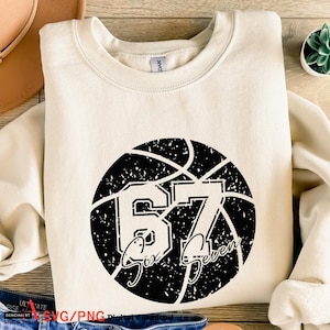 Puede incluir: Sudadera color crema con un gráfico de baloncesto negro desgastado con el número 67 y las palabras "Six Seven". La sudadera está hecha de un material suave y tiene cuello redondo.