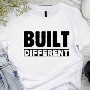 以下が含まれることがあります： 「BUILT DIFFERENT」の文字が黒い太字でプリントされた白いTシャツ。 「DIFFERENT」の文字は黒い長方形の中にあります。 Tシャツは明るい木目調の表面に、青いジーンズと一緒に置かれています。