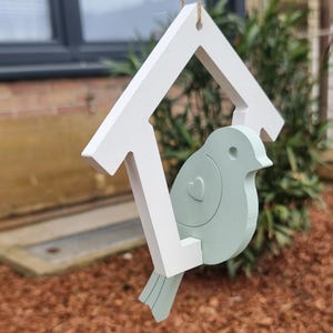 Könnte beinhalten: Ein weißes Vogelhaus aus Holz mit einem hellgrünen Vogel mit einem Herzen auf der Brust, das an einer Schnur hängt.