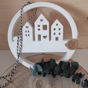 Könnte beinhalten: Weiße Holzdekoration mit drei stilisierten Häusern in einem kreisförmigen Rahmen. Die Häuser haben Herz-, Quadrat- und Sternausschnitte. Ein schwarz-weißes Seil hängt von oben. Ein kleines Etikett und Grünzeug sind ebenfalls vorhanden.