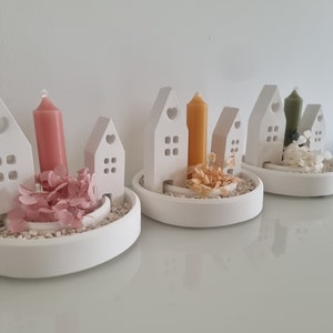 May include: White ceramic candle holders shaped like houses, each holding a different colored candle. The holders are arranged in a row, with pink, yellow, and green candles. Decorative dried flowers and small stones are also included.