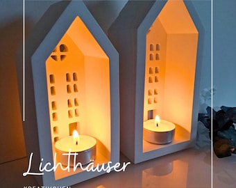 May include: Two white ceramic house-shaped candle holders with cut-out windows and doors. Each house contains a lit tea light, casting a warm glow. The word "Lichthäuser" is written in cursive on the front.
