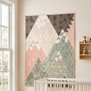 May include: A rectangular wall quilt featuring a geometric mountain landscape design composed of triangular fabric patches in shades of dusty pink, sage green, charcoal grey, and cream, finished with a thin pink border and quilted texture.