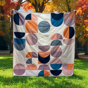 May include: A handmade quilt with a geometric design of half-circles and full circles in shades of navy, orange, pink, and purple. The quilt is displayed outdoors on a grassy lawn, with autumn foliage in the background. The quilt is approximately 70 inches by 70 inches.