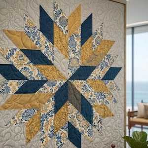 Op de afbeelding: Een ingelijste quilt met een sterrenpatroon. De quilt heeft een patroon van marineblauwe, gouden en cr&egrave;mekleurige bloemenstofstukken. De achtergrond is lichtgrijs met een gewatteerd patroon. De quilt wordt tentoongesteld in een kamer met uitzicht op de oceaan.