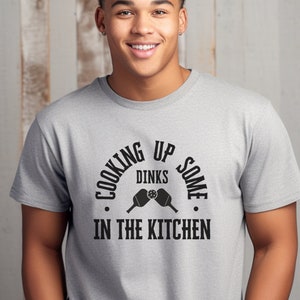 Pode incluir: Uma camiseta cinza com um gráfico preto que diz "Cooking Up Some Dinks In The Kitchen". O gráfico apresenta um par de espátulas cruzadas. A camiseta está disponível na cor Cinza Esportivo.