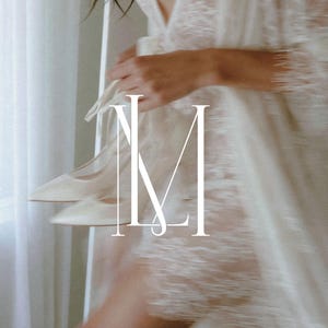 May include: A pair of white pointed-toe high-heeled shoes held alongside a white lace robe with long sleeves and intricate floral patterns. The letters LM are overlaid in white in the centre of the image, partially obscuring the shoes and robe.